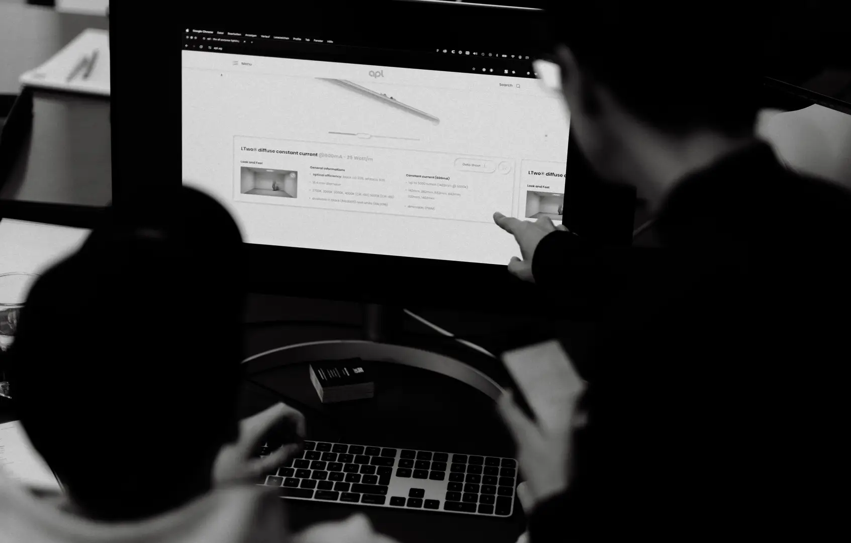Schwarz-weißes Foto zweier Mitarbeiter von hinten, die gemeinsam an einem Monitor eine Produktdetailseite mit technischen Zeichnungen begutachten, einer zeigt mit dem Finger auf den Bildschirm.