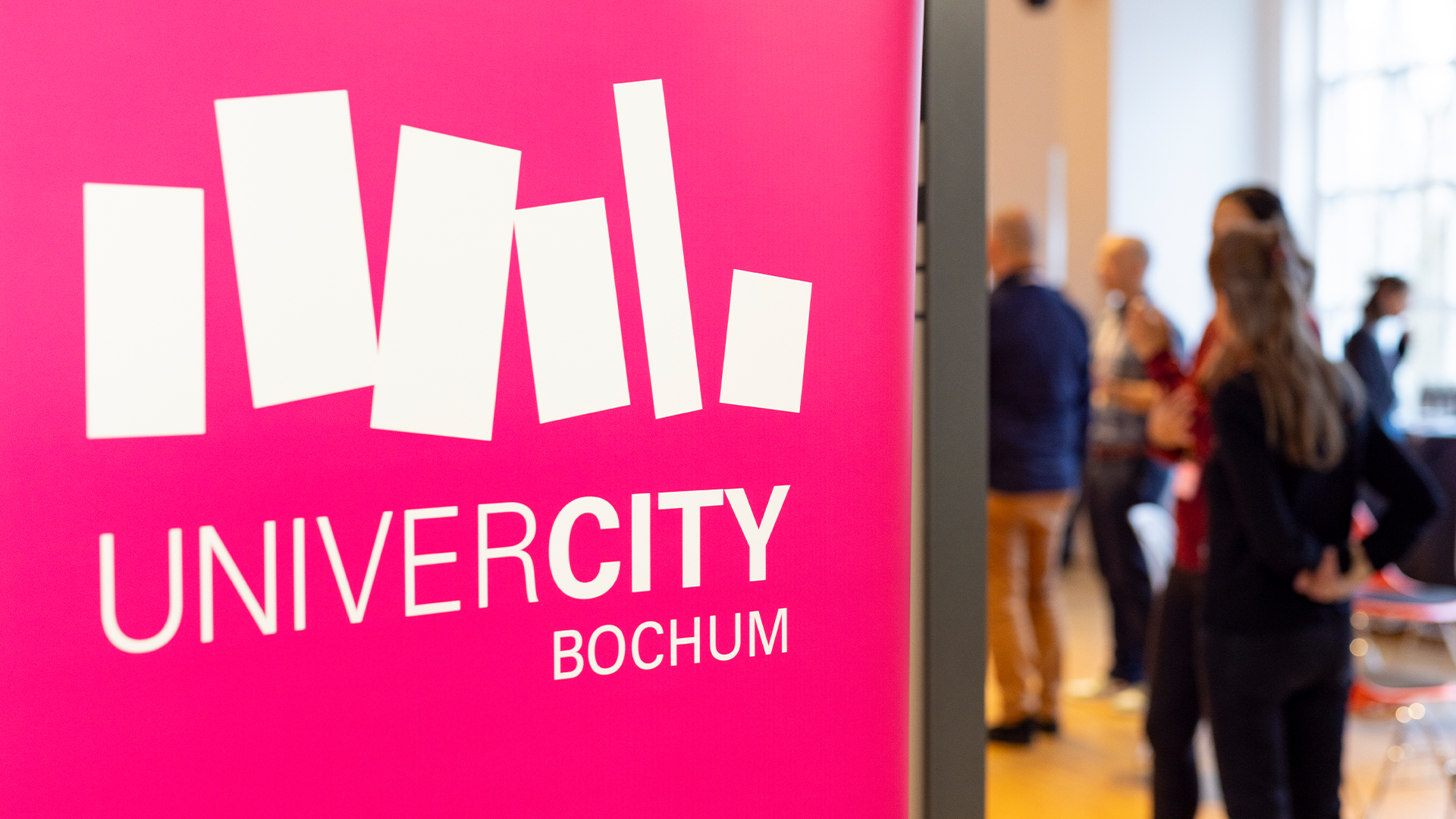 Pinkfarbener Aufsteller mit UniverCity-Bochum-Logo, dahinter Teilnehmende im Gespräch