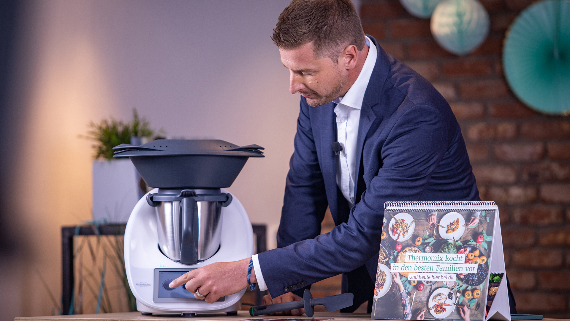 Moderator im Anzug bedient einen Thermomix, daneben ein Tischaufsteller mit Thermomix-Motiv