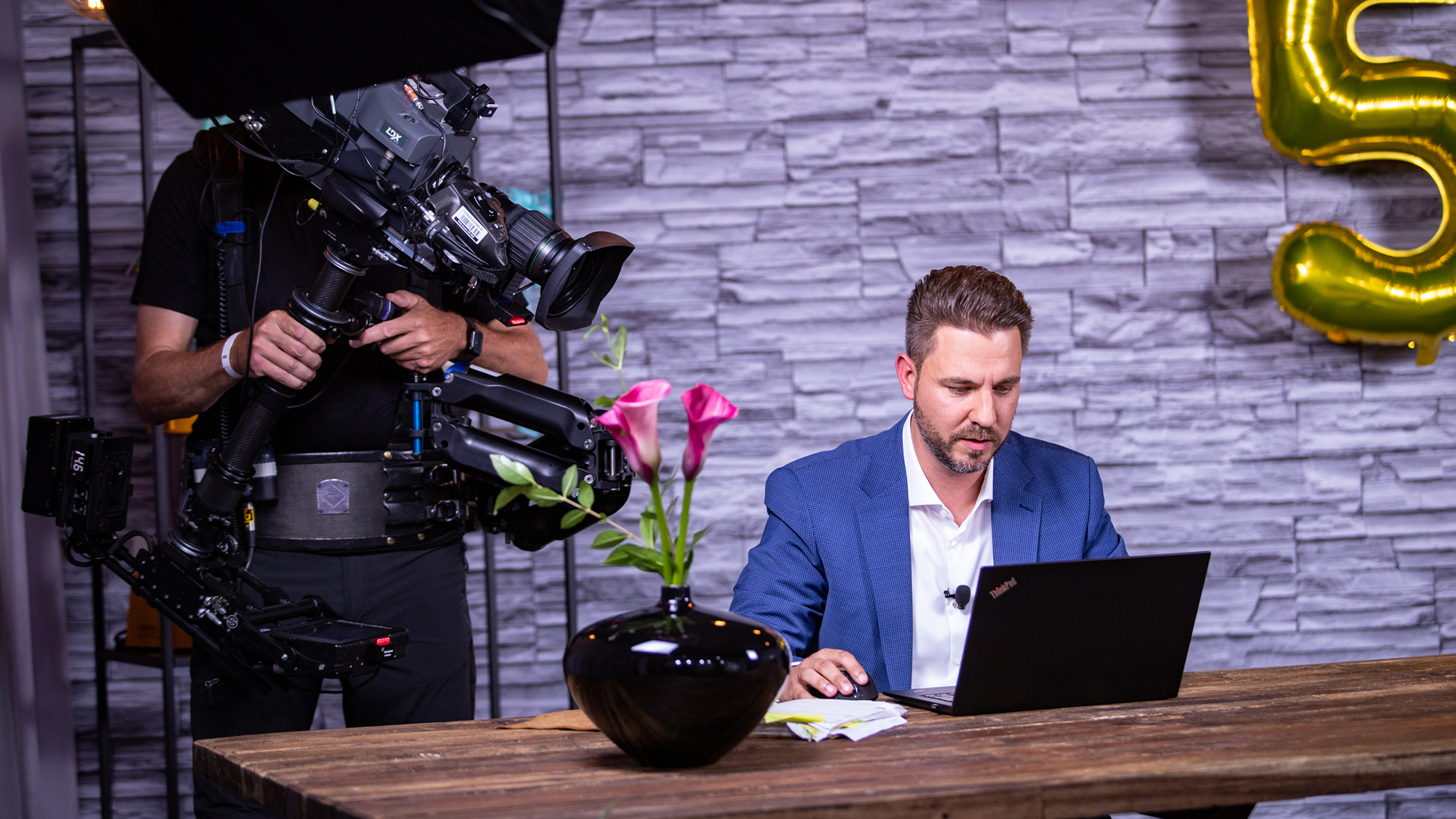 Moderator am Laptop an einem Holztisch, links ein Kameramann mit Steadicam