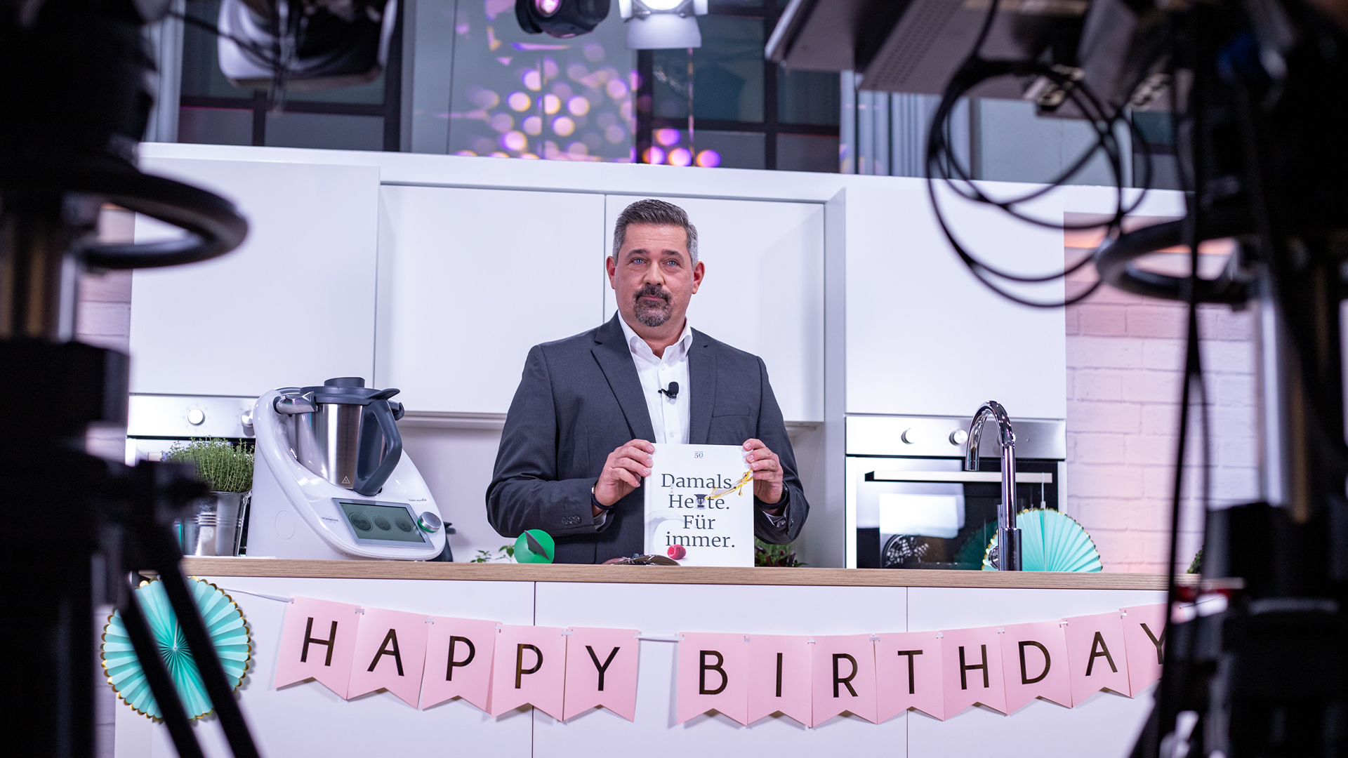 Mann in Küchenkulisse hält ein Jubiläumsbuch, davor Thermomix und Happy-Birthday-Girlande